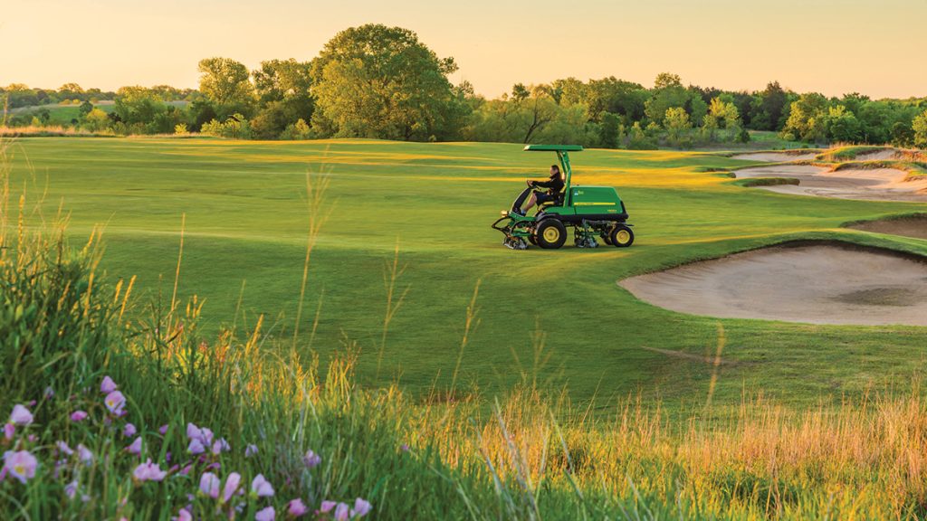 A photo of a John Deere Fairway Mower 7500A taken from a distance on a golf course. The mower is in the background as its' being driven around to cut the grass of the fairway. There are sandpits behind the mower to the right of the photo. The foreground is a small hill, covered in pink flowers and slightly taller grass. In the photo you can tell the sun is busy setting. Past the mower you can see some trees and bushes.