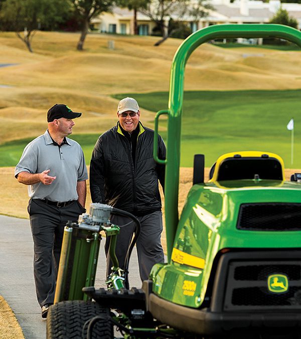 A photo of a John Deere Fairway Mower 7500A taken from the front as a close-up. The focus of the photo is on the two men behind the mower as they walk down a pathway on the golf course while talking. One man is gesturing with his hand as the other laughs.  Past the men you can see some small rolling hills that form part of the golf course and way in the background is a building, probably the club house of the course.