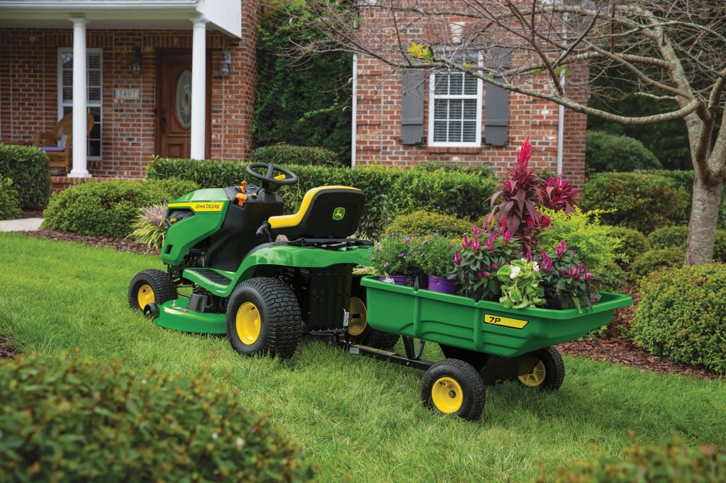 A photo of a John Deere S100 Ride-On Mower with an attachment. It is standing in front of a garden on the grass on the yard of a big house. The attachment is a type of trolley loaded with plants