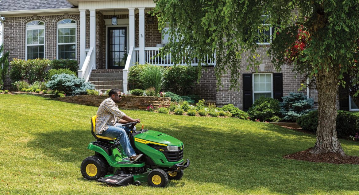 A photo of a John Deere S140 Ride-On Mower as a person drives it on a front yard to mow the lawn in front of a big house.