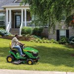 A photo of a John Deere S140 Ride-On Mower as a person drives it on a front yard to mow the lawn in front of a big house.