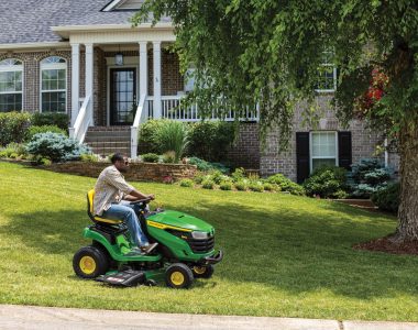 A photo of a John Deere S140 Ride-On Mower as a person drives it on a front yard to mow the lawn in front of a big house.