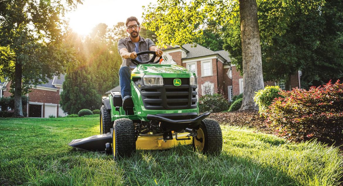 A photo of a John Deere S160 Ride-On Mower driving towards the camera with a man driving it and cutting the lawn.