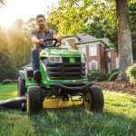 A photo of a John Deere S160 Ride-On Mower driving towards the camera with a man driving it and cutting the lawn.