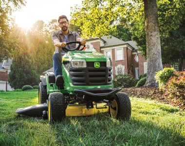 A photo of a John Deere S160 Ride-On Mower driving towards the camera with a man driving it and cutting the lawn.