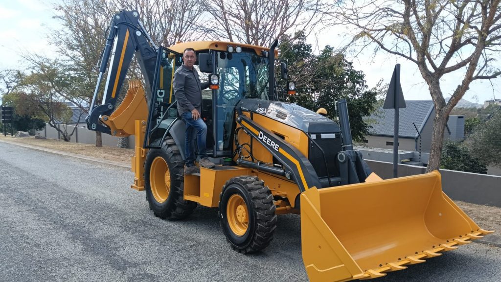 A photo of a John Deere TLB on a gravel road in what looks like a residential area with houses and trees in the background. At the door to the cab of the TLB, George Nel stands, about to climb in, looking at the camera.