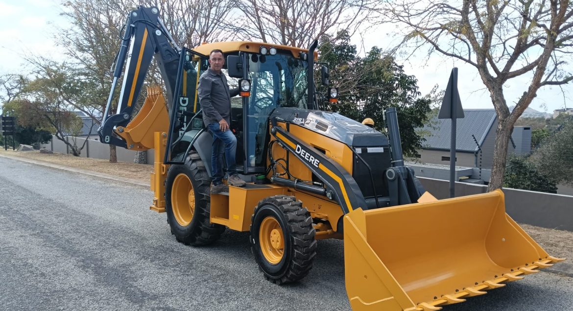 A photo of a John Deere TLB on a gravel road in what looks like a residential area with houses and trees in the background. At the door to the cab of the TLB, George Nel stands, about to climb in, smiling at the camera.