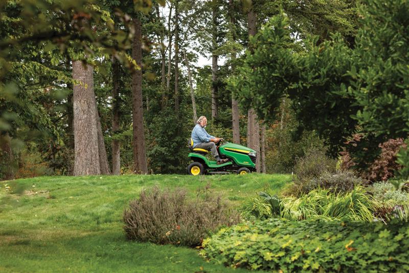 A photo of a John Deere Ride-On Mower in the distance on a slight uphill as a person drives it to cut the grass. There are tress in the background and a garden in the foreground.