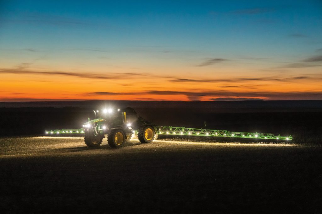 A photo of a John Deere 612R Self-Propelled Sprayer as it sprays during the Night in the field with the light on as the sun is setting in the background