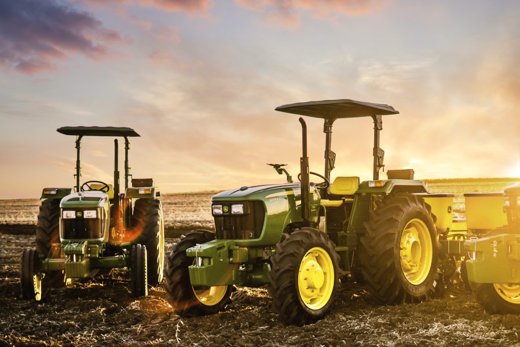 A photo of a 5055E and 5075E John Deere tractors as they stand in a field with the sun setting behind.
