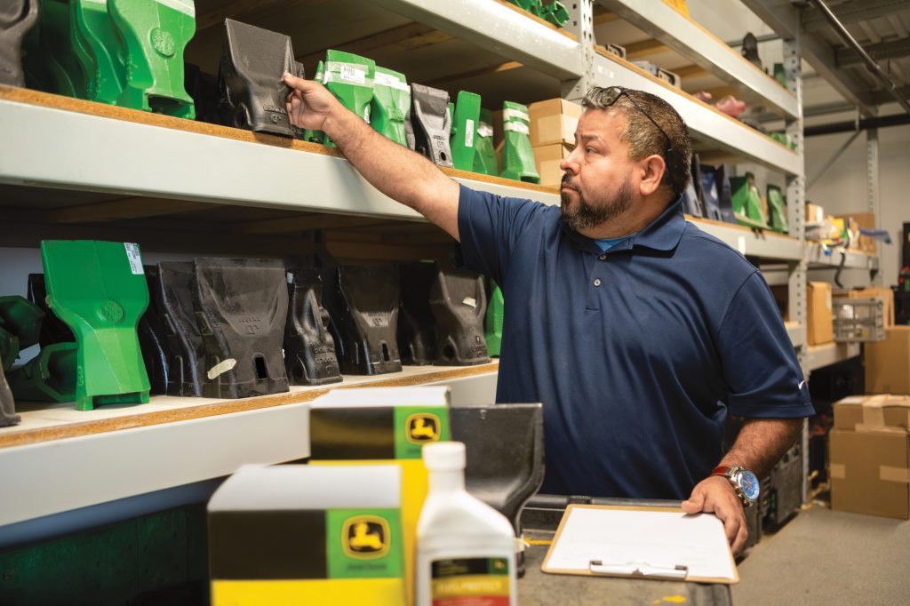 A parts salesman reaching onto a shelf to pull a genuine John Deere part from a shelf lined with differnt John Deere parts. In the foreground you can make out a blurry shape of genuine John Deere service kits and other bottles.