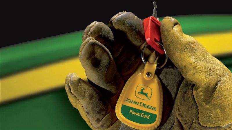 A key of a John Deere machine with a keychain that reads PowerGard in a gloved hand of technician with a John Deere tractor in the background