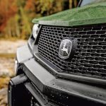 A photo of the nose of the John Deere XUV 875M Diesel parked on farm terrain. The close-up shows the John Deere logo.