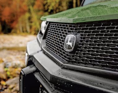 A photo of the nose of the John Deere XUV 875M Diesel parked on farm terrain. The close-up shows the John Deere logo.