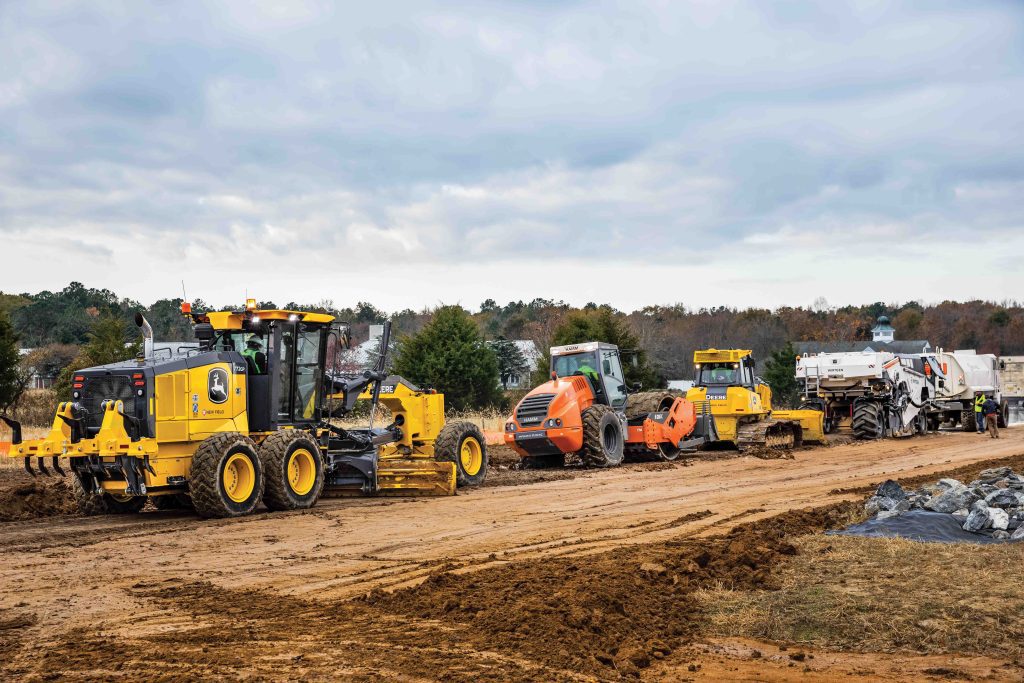 John Deere road construction equipment and Wirtgen machines, including a motor grader and other machines, working on a roadbuilding project.